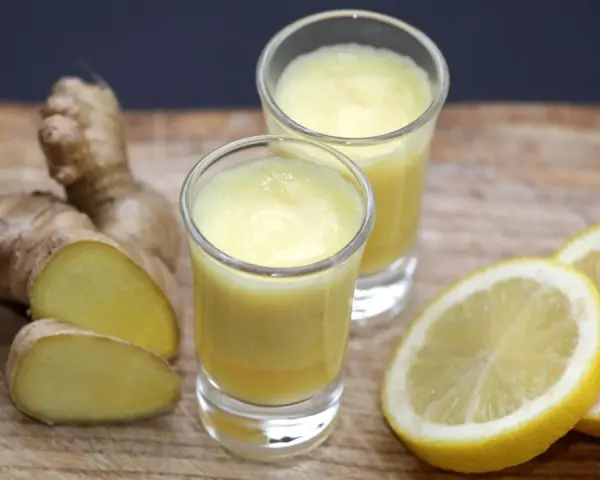 Zwei Gläser mit leuchtend gelbem Ingwer Shot, daneben frischer Ingwer und Zitronenscheiben auf Holzbrett.