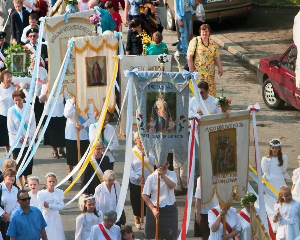 Procesja z banerami religijnymi, w tym z napisem "Boże Ciało 26". Ludzie niosą obrazy i kwiaty.