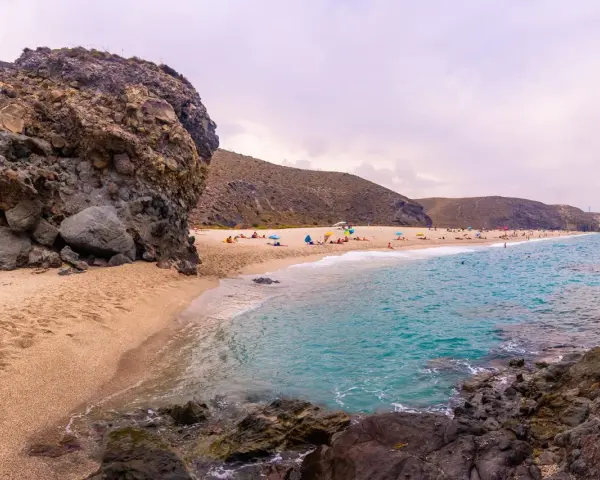 Plaża w Cabo de Gata z turkusową wodą, piaszczystym brzegiem i skalistymi formacjami. Ludzie odpoczywają pod parasolami.
