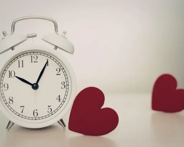 Biały budzik pokazuje 10:08. Obok niego leżą dwa czerwone serca. 0202 znaczenie: czas na miłość.