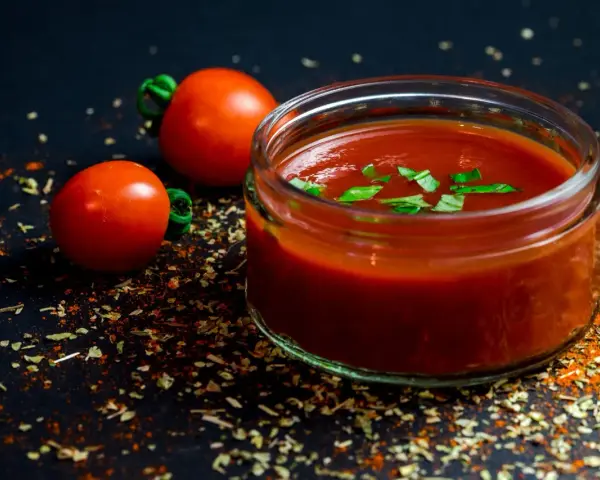 Un vasetto di ketchup fatto in casa, guarnito con basilico fresco, accanto a pomodori maturi e spezie su sfondo scuro.