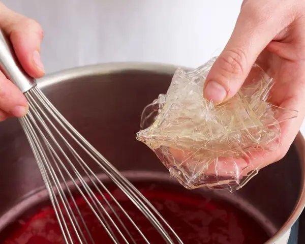 Hände bereiten Gelatine-Blätter für die Zubereitung vor, indem sie sie in eine rote Flüssigkeit in einem Topf geben, während ein Schneebesen bereitsteht.