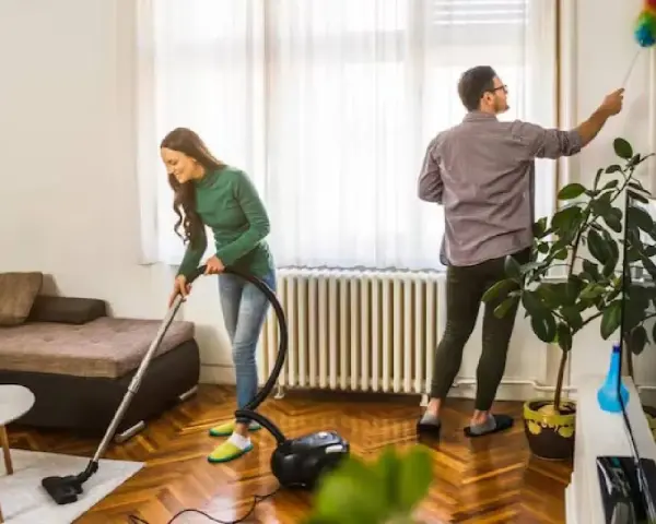 Pareja colaborando para purificar casa. Ella aspira el suelo, él quita el polvo de las paredes.
