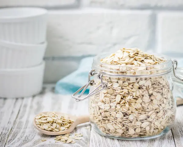 Ein Glas voller haferflocken gemahlen, bereit für ein gesundes Frühstück. Daneben ein Holzlöffel mit weiteren Flocken.