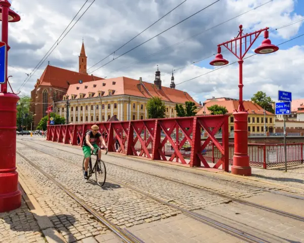 Mężczyzna na rowerze jedzie po brukowanej drodze rowerowej z torami tramwajowymi, w tle zabytkowe budynki i czerwony most.