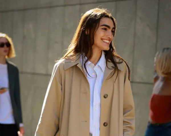 Donna sorridente con capelli lunghi e castani indossa un trench beige aperto su una camicia a righe azzurre. Perfetto per ogni occasione, anche se posso mettere il trench di sera.