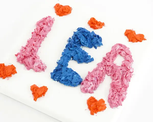 Comment faire du relief sur un tableau : des lettres "LÉA" en papier froissé rose et bleu, entourées de cœurs orange sur une toile blanche.