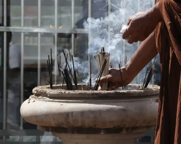 Manos monje en túnica marrón colocando varillas de incienso en un cuenco de piedra, creando humo para limpiar el ambiente.