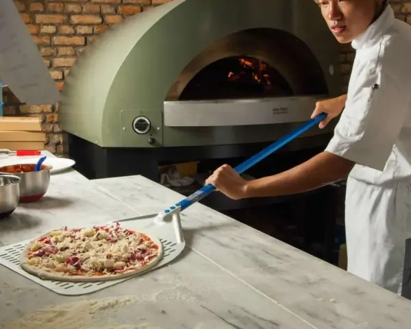 Pizza cruda en pala, lista para horno de leña con chef al fondo.