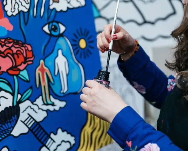 Une artiste, dans son métier artistique, trempe son pinceau dans l'encre pour peindre une toile aux motifs surréalistes : une fleur, un œil, des silhouettes.