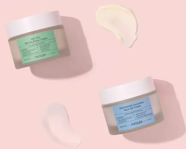 Due barattoli di crema viso Meisani, uno con niacinamide e burro, l'altro con niacinamide e cetriolo, su sfondo rosa. Perfetti per proteggere la pelle dal sole.