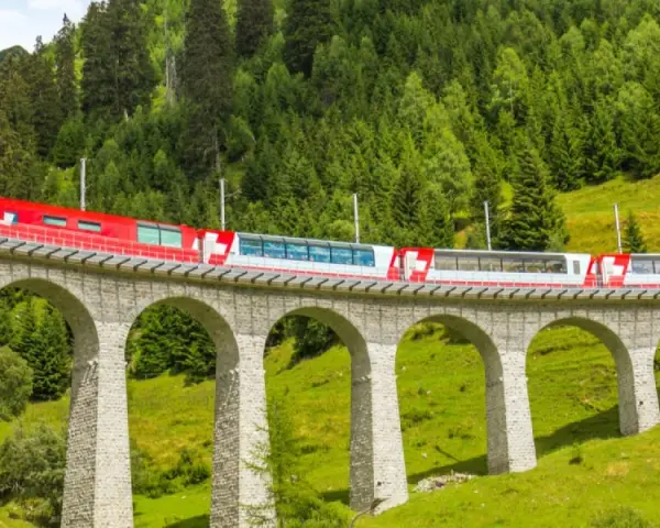 A red and white train travels across a stone viaduct, winding through a lush green valley with a dense forest on the mountainside. This scenic route is part of the Amsterdam to Basel train journey.