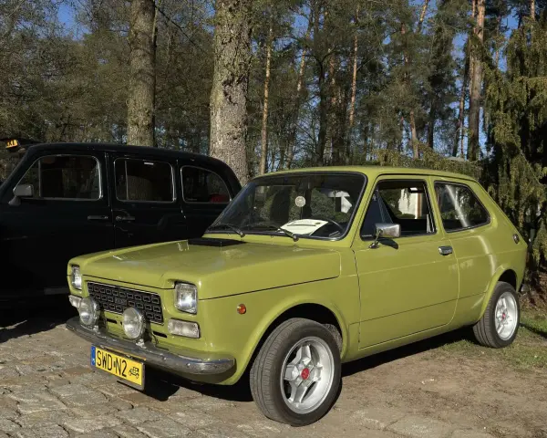 Zielony fiat 127 z lat 70. stoi obok czarnej taksówki na tle drzew.