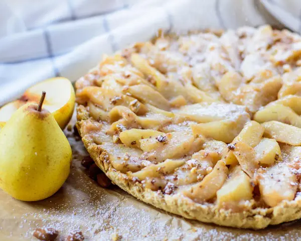 Délicieuse tarte aux poires, saupoudrée de sucre glace, avec des amandes et des poires fraîches à côté.