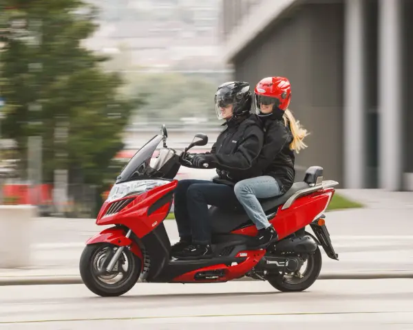 Para jadąca na czerwonym skuterze 125, ubrana w kurtki i kaski, cieszy się jazdą.