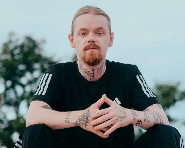 Mężczyzna z rudą brodą i blond włosami, ubrany w czarną koszulkę z trzema paskami. Na szyi i rękach widoczne tatuaże, w tym klucz i napis "kuqe 2115".