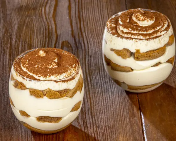 Due bicchieri di tiramisù senza uova, stratificati con crema e savoiardi, spolverati di cacao su un tavolo di legno.