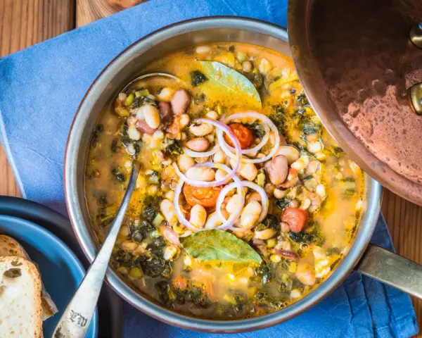 Zuppa ricca con fagioli, verdure e spezie, servita in una pentola di rame. Un piatto confortante di acqua cotta, guarnito con cipolla rossa e alloro.