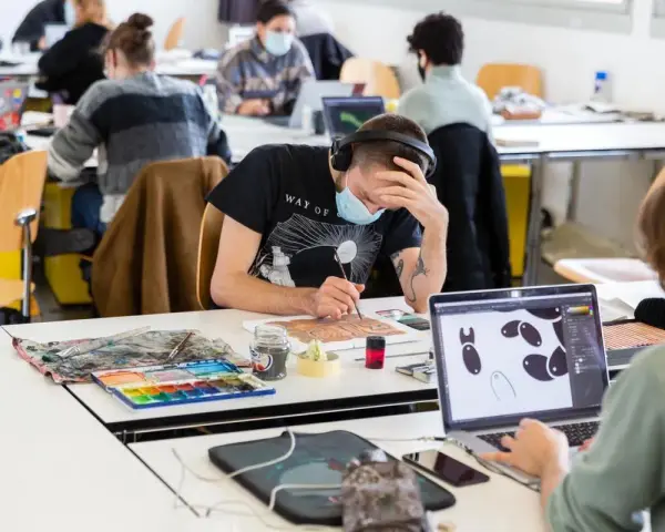 Des étudiants concentrés dans une école d'art, certains peignent, d'autres travaillent sur ordinateur. L'ambiance est studieuse.