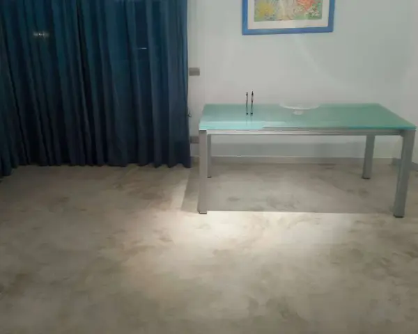 Un tavolo moderno con piano in vetro poggia su un **pavimento in cemento grezzo** dall'aspetto materico, con tende blu sullo sfondo.