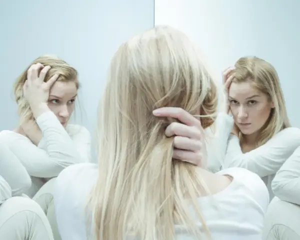 Blondynka w białej bluzie, zmagająca się z kwetaplex, widzi swoje odbicia w lustrze, każde z innym wyrazem twarzy.