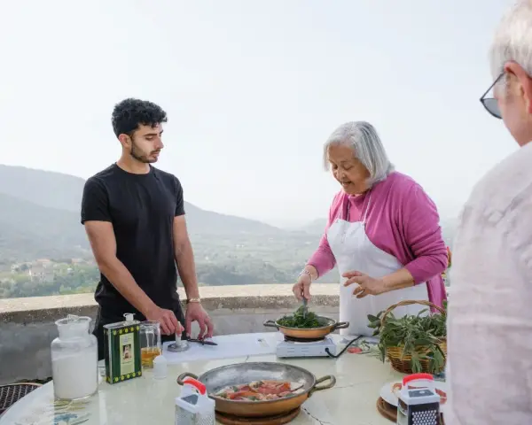 An older woman in an apron cooks greens while a young man and an older man stand nearby on a terrace with a scenic view.