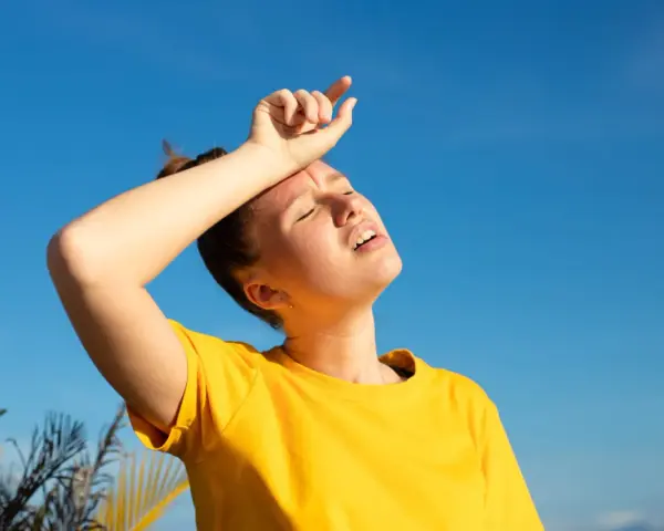 Une jeune femme en t-shirt jaune, le visage marqué par la chaleur, essuie son front sous un ciel bleu intense. La durée d'une insolation est palpable.