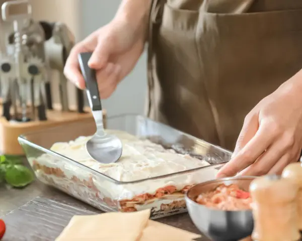 Osoba nakłada sos na warstwy makaronu i mięsa, przygotowując do pieca lasagne.