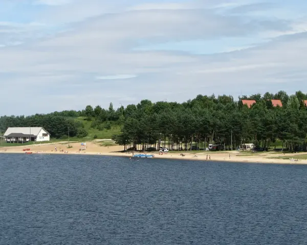 Plaża nad jeziorem Chańcza tętni życiem. Ludzie odpoczywają na piasku, kąpią się w wodzie, a w tle widać domki i las.