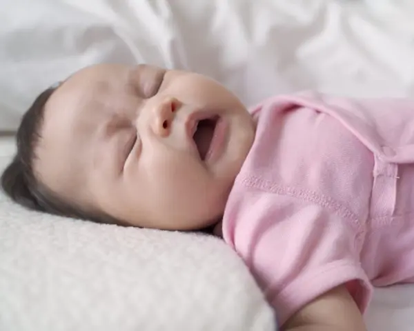 Ein süßes neugeborenes Baby in rosa Strampler niest. Die Augen sind geschlossen, der Mund leicht geöffnet.