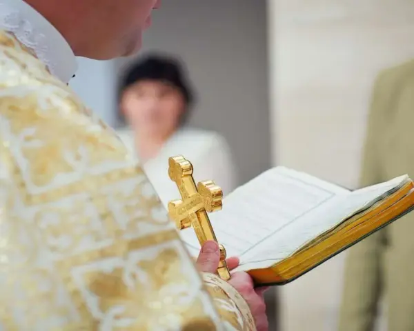 Ksiądz w złotej szacie trzyma krzyż i księgę podczas uroczystości. To ważny moment, kiedy odbywa się bierzmowanie.