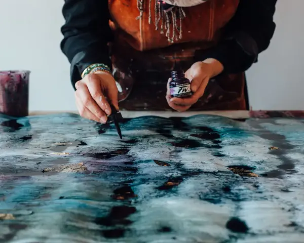 Une artiste crée une **aquarelle sur toile** aux teintes bleues et dorées, ajoutant des touches d'encre avec un compte-gouttes.