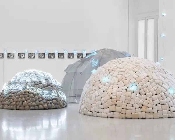 Installation d'art povera : dômes en pierres et en tissu, ornés de néons bleus, sous une structure transparente.