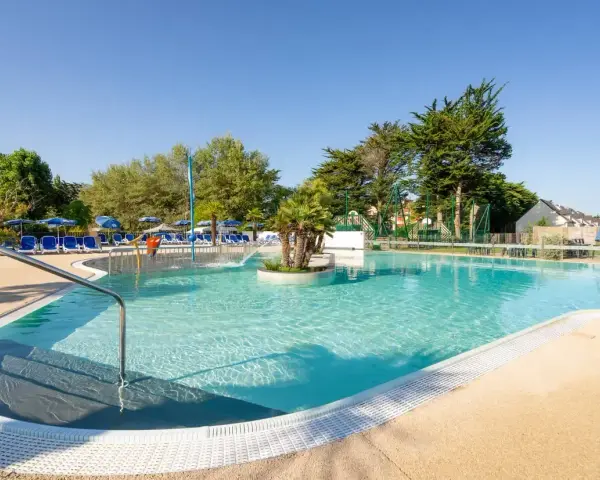 Piscine extérieure avec chaises longues bleues, parasols et végétation luxuriante sous un ciel bleu clair.
