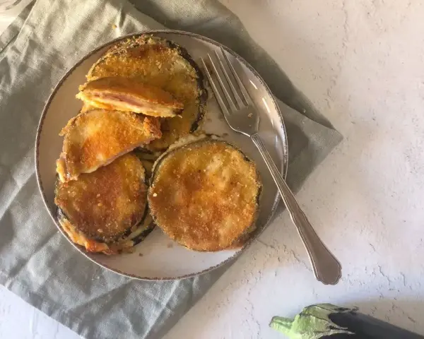 Cordon bleu di melanzane impanati e fritti, ripieni di prosciutto e formaggio filante, serviti su un piatto con una forchetta.