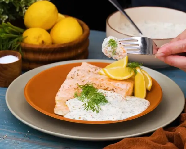 En bit lax med citronskivor och dill, toppad med en krämig romsås till lax. En gaffel med romsås lyfts från skålen.