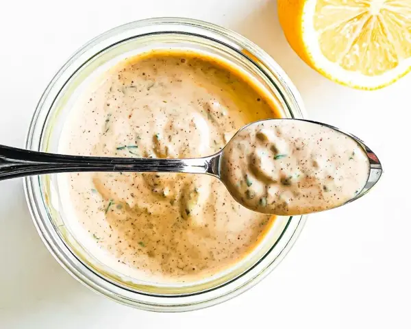En sked krämig remouladsås med örter lyfts ur en burk. Bredvid ligger en halv citron. Perfekt för ditt remouladsås recept!