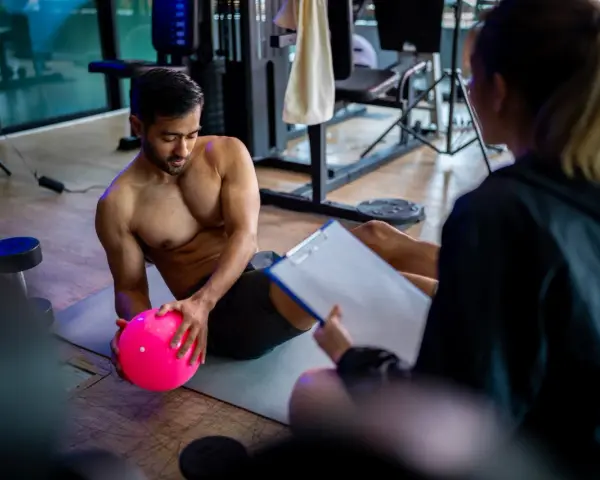 Man med rosa boll på gymmatta, tränare med clipboard. De följer sitt gymprogram.