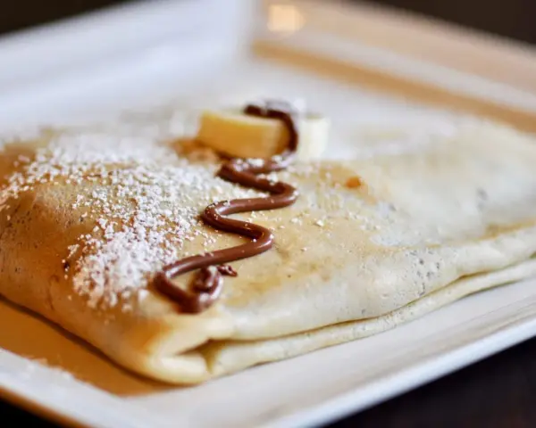 Crêpe pliée, saupoudrée de sucre glace, décorée de chocolat fondu et d'une rondelle de banane. La meilleure recette de pâte à crêpes pour un délice !