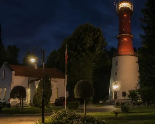 Wieczorna iluminacja latarni morskiej Rozewie. Obok niej biały budynek i ceglany komin.