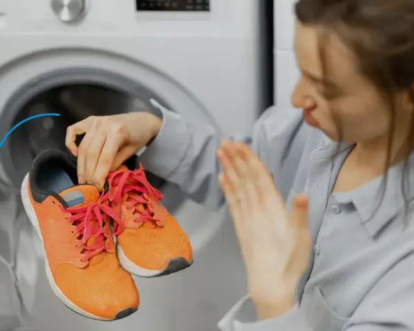 Woman holding smelly orange sneakers near an open washing machine.