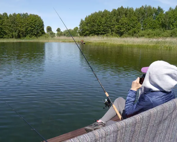 Osoba siedzi na pomoście i łowi ryby na jeziorze Jagodne. W tle las i trzciny.