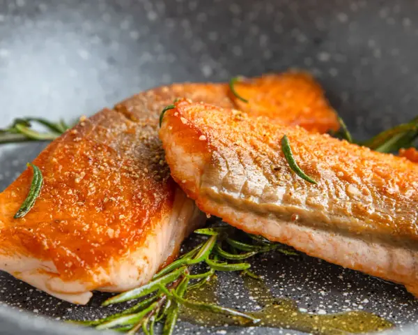 Zwei saftige Lachsfilets mit knuspriger Haut, perfekt gebraten, liegen in einer Pfanne mit Rosmarin und Öl.