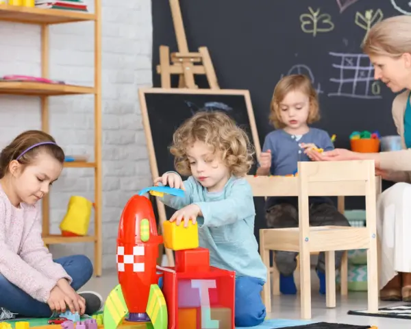 Dzieci budują rakietę z klocków, a opiekunka czuwa. Ciekawe, ile zarabia opiekunka w żłobku za taką pracę.