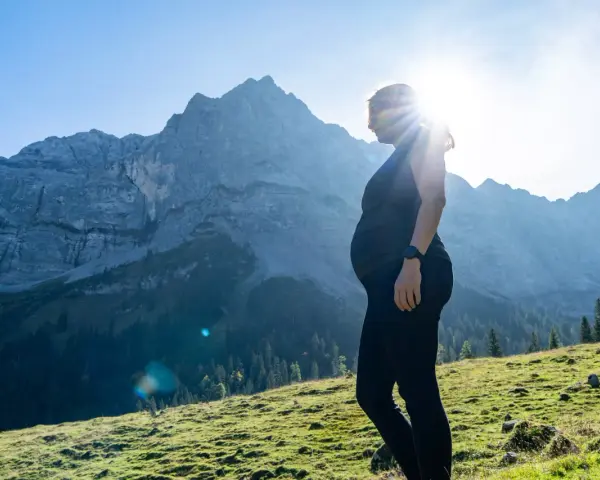 Frau in der Schwangerschaft genießt das Wandern in den Bergen. Die Sonne scheint hinter ihr, während sie auf eine majestätische Bergkulisse blickt.