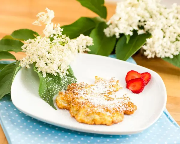 Fiori di sambuco fritti, spolverati di zucchero a velo, serviti su un piatto bianco con fragole fresche e decorati con fiori di sambuco.