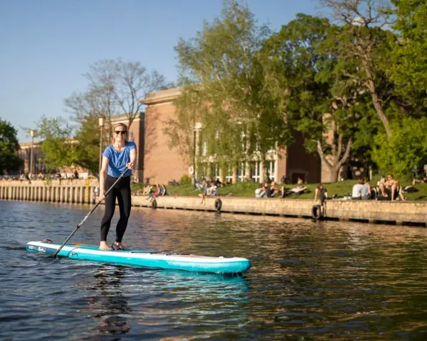 Frau genießt Stand Up Paddling auf einem Fluss, während Menschen am Ufer entspannen.