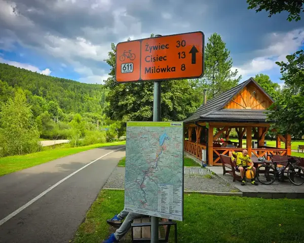 Znak szlaku rowerowego 611 Velo Soła wskazuje kierunki. Obok mapa i wiata z rowerzystą.