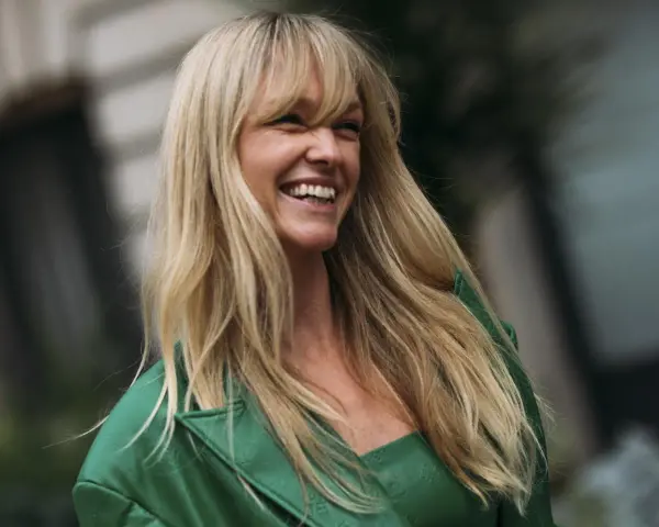 Frau mit langen blonden Haaren und Pony, die lacht und ein grünes Jackett trägt.