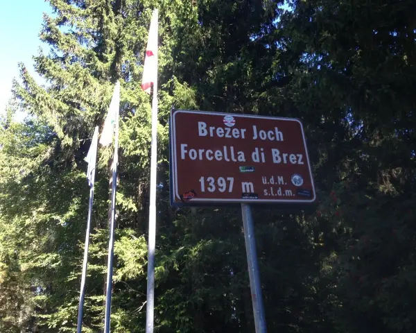 Cartello indicante "Forcella di Brez" a 1397 m, con bandiere e alberi sullo sfondo.